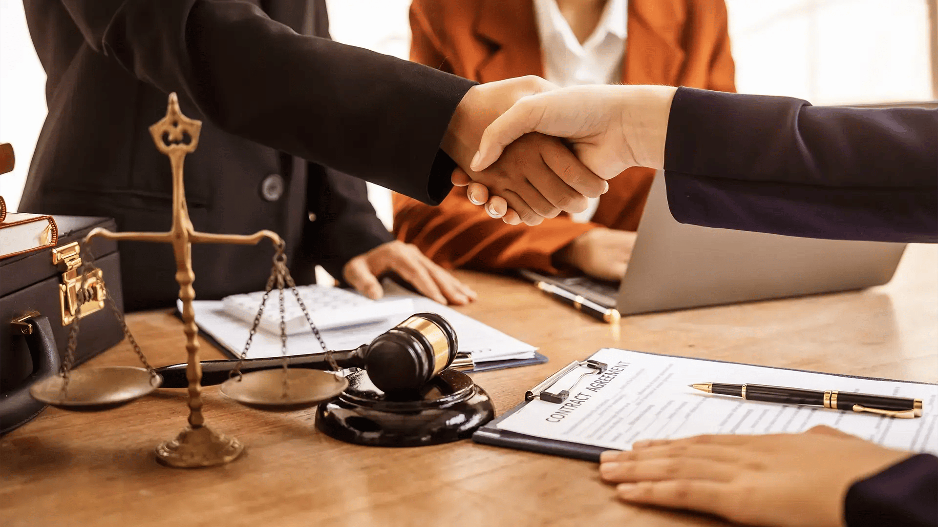 Dos personas estrechando la mano sobre una mesa con mazo judicial, balanza de justicia y contrato, en reunión legal.